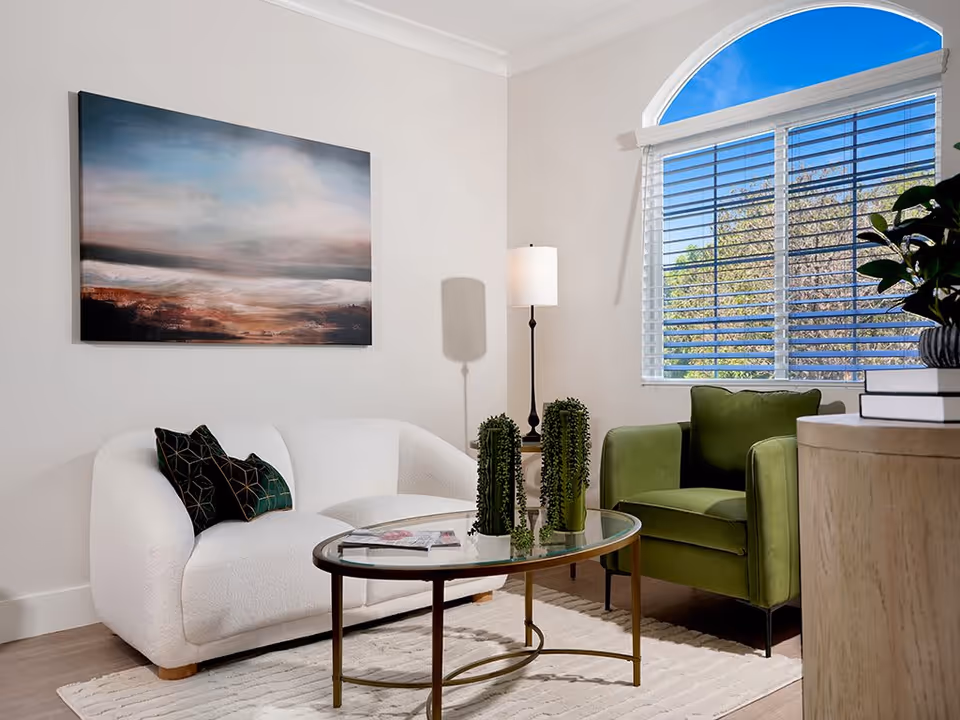 A cozy living room with a white curved sofa adorned with two dark green pillows, a green armchair, a glass coffee table with two small potted plants and magazines, a floor lamp, a large window with white blinds, and a landscape painting on the wall.