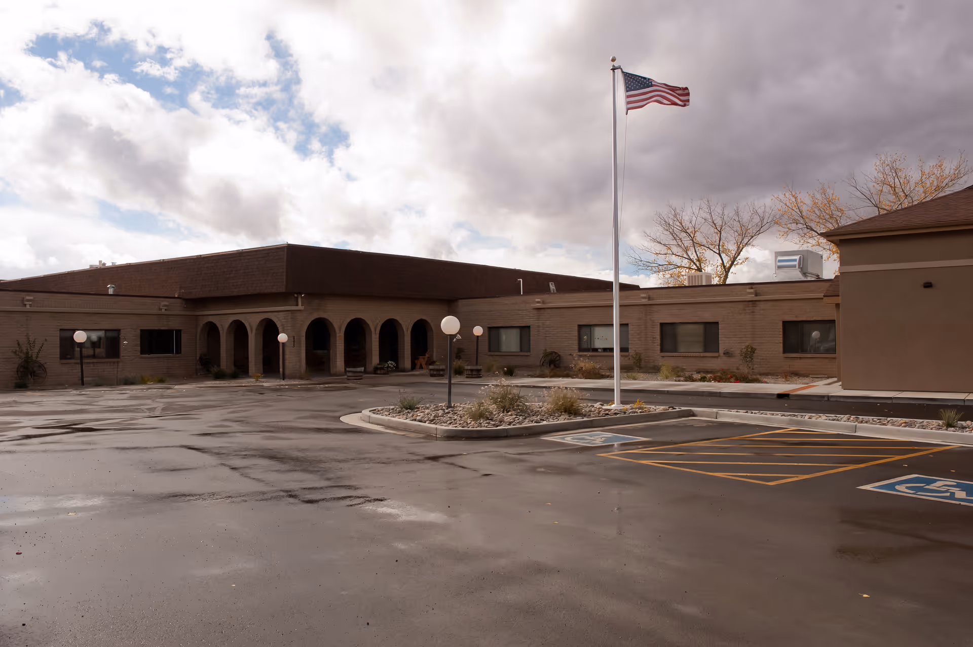 Exterior view of Colorow Care Center building with a wet parking lot in front, an American flag on a flagpole, several round street lamps, and cloudy sky overhead.