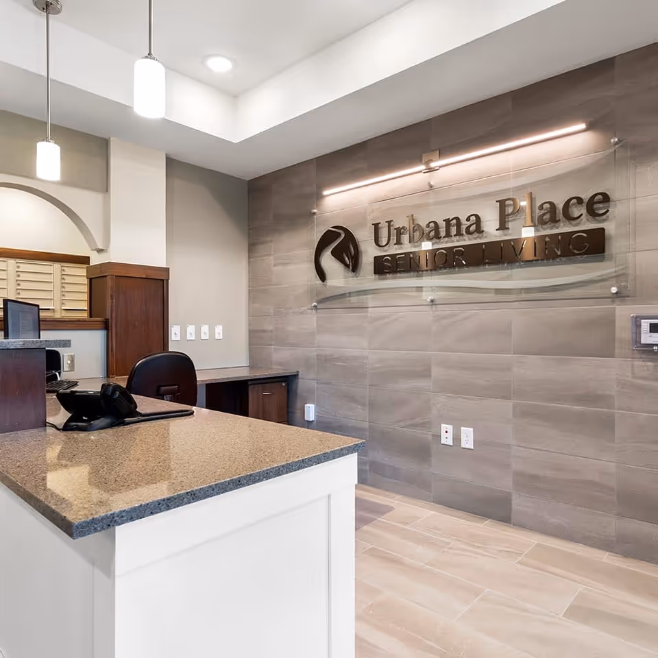 Reception area of Urbana Place Senior Living featuring a modern desk with a phone and computer, a chair, and a wall with the Urbana Place Senior Living logo mounted on it. The space is well-lit with pendant lights and has a clean, professional appearance.