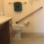 A bathroom with a white toilet, a grab bar on the wall beside the toilet, a toilet paper holder with a roll of toilet paper, a towel rack with a green towel and a brown washcloth, and a wooden cabinet with a white countertop.