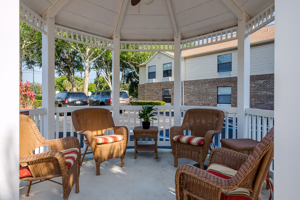 Outdoor gazebo area with four wicker chairs featuring red and beige striped cushions arranged around a small wicker table with a potted plant. The gazebo has white railings and a white roof. In the background, there are parked cars, trees, and a two-story building with brick and beige siding.