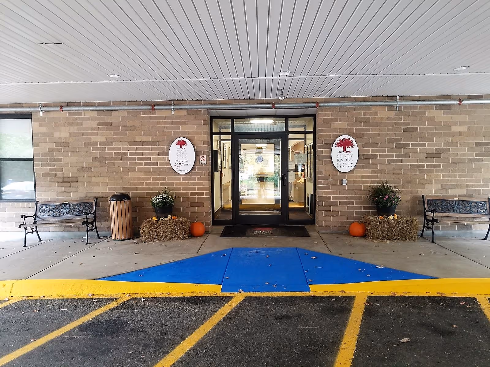Entrance to Shady Knoll Health Center featuring double glass doors framed by brick walls. There are two benches on either side of the entrance, a trash can, and decorative hay bales with pumpkins and potted plants. The area is covered by a white ceiling with recessed lighting, and a blue and yellow painted curb is visible in front of the entrance.