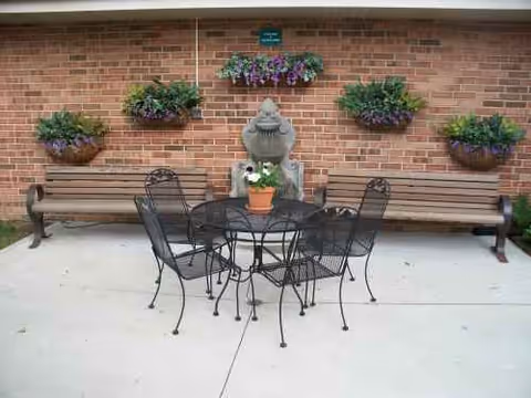 Outdoor patio with metal table and chairs, benches, and flower pots against a brick wall.