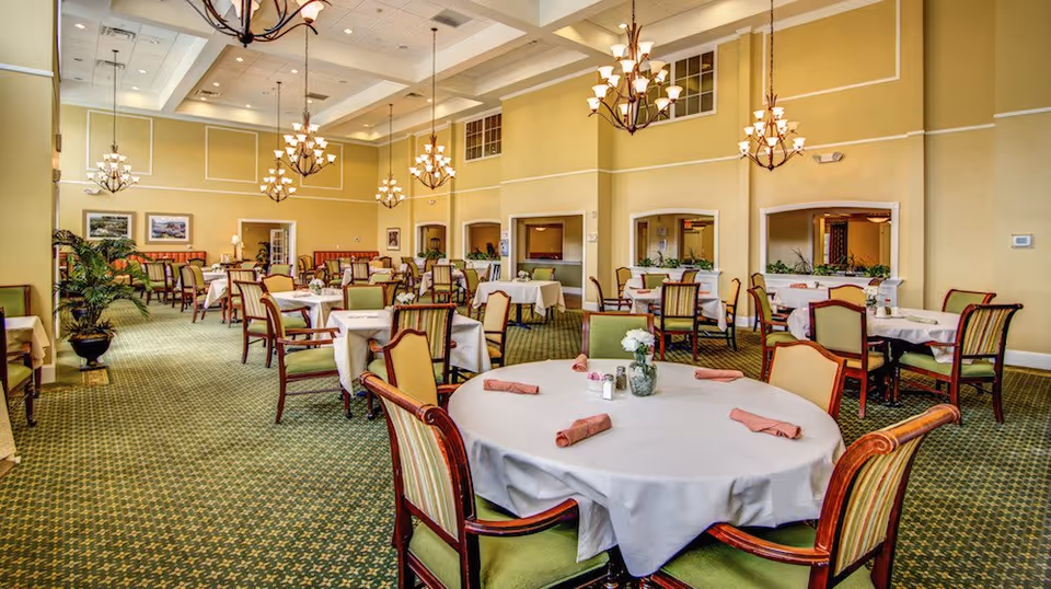Spacious dining room with multiple round and rectangular tables covered with white tablecloths, each set with folded napkins and small floral centerpieces. The room features green patterned carpet, high ceilings with multiple chandeliers, and light yellow walls with white trim. Several chairs with green and beige upholstery surround the tables, and there are plants and framed pictures along the walls.