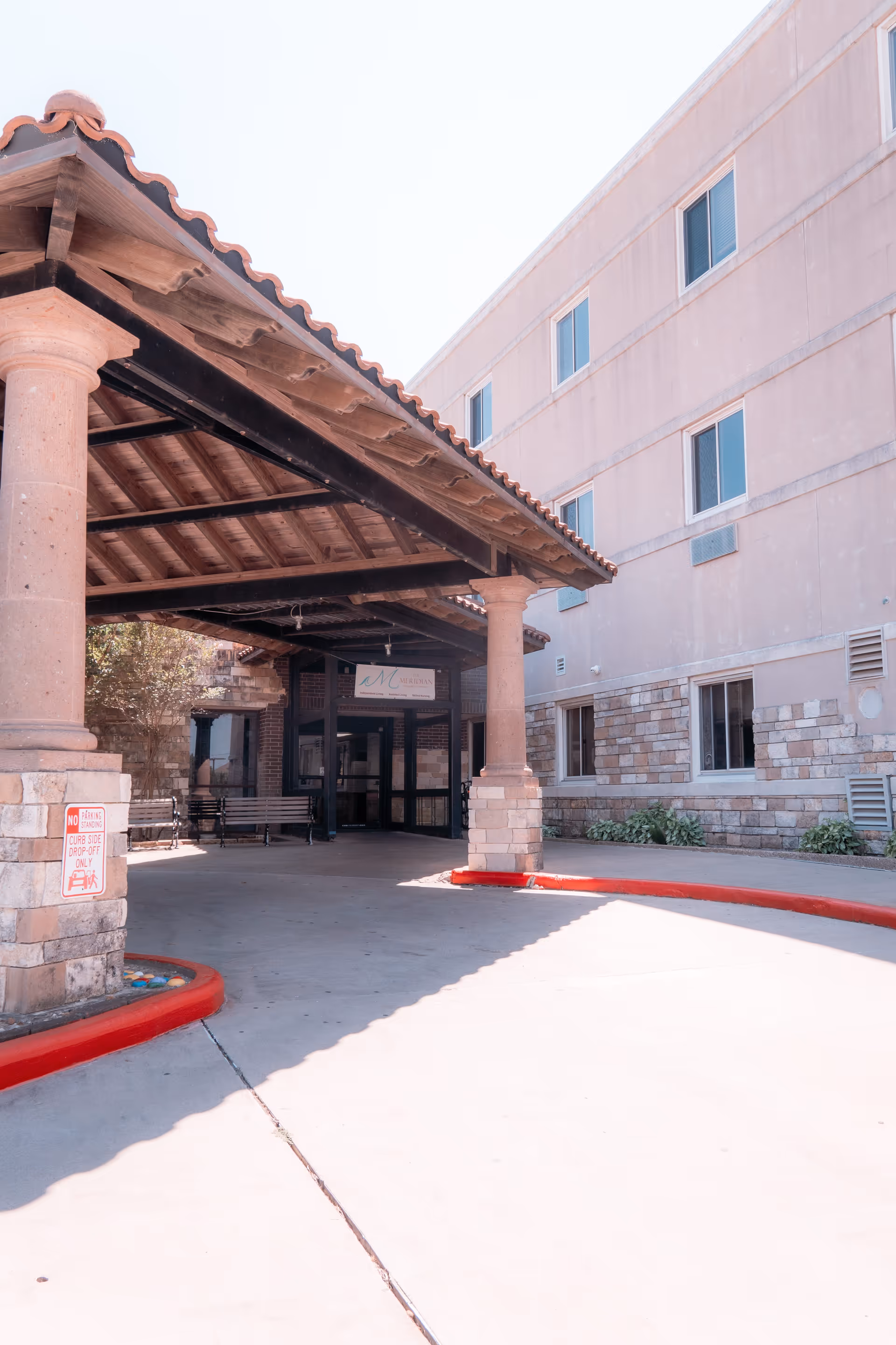 Front covered entrance (porte-cochere) of a multi-story senior living building with stone pillars and a curved driveway.