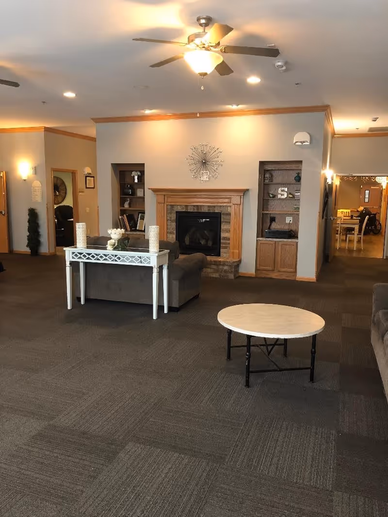 A cozy living room area in an assisted living facility featuring a ceiling fan with light, a fireplace with a wooden mantel, built-in shelves on either side of the fireplace, a gray couch with a white console table behind it, and a round coffee table in the foreground. The room has carpeted flooring and warm lighting, with a hallway visible in the background leading to a dining area.
