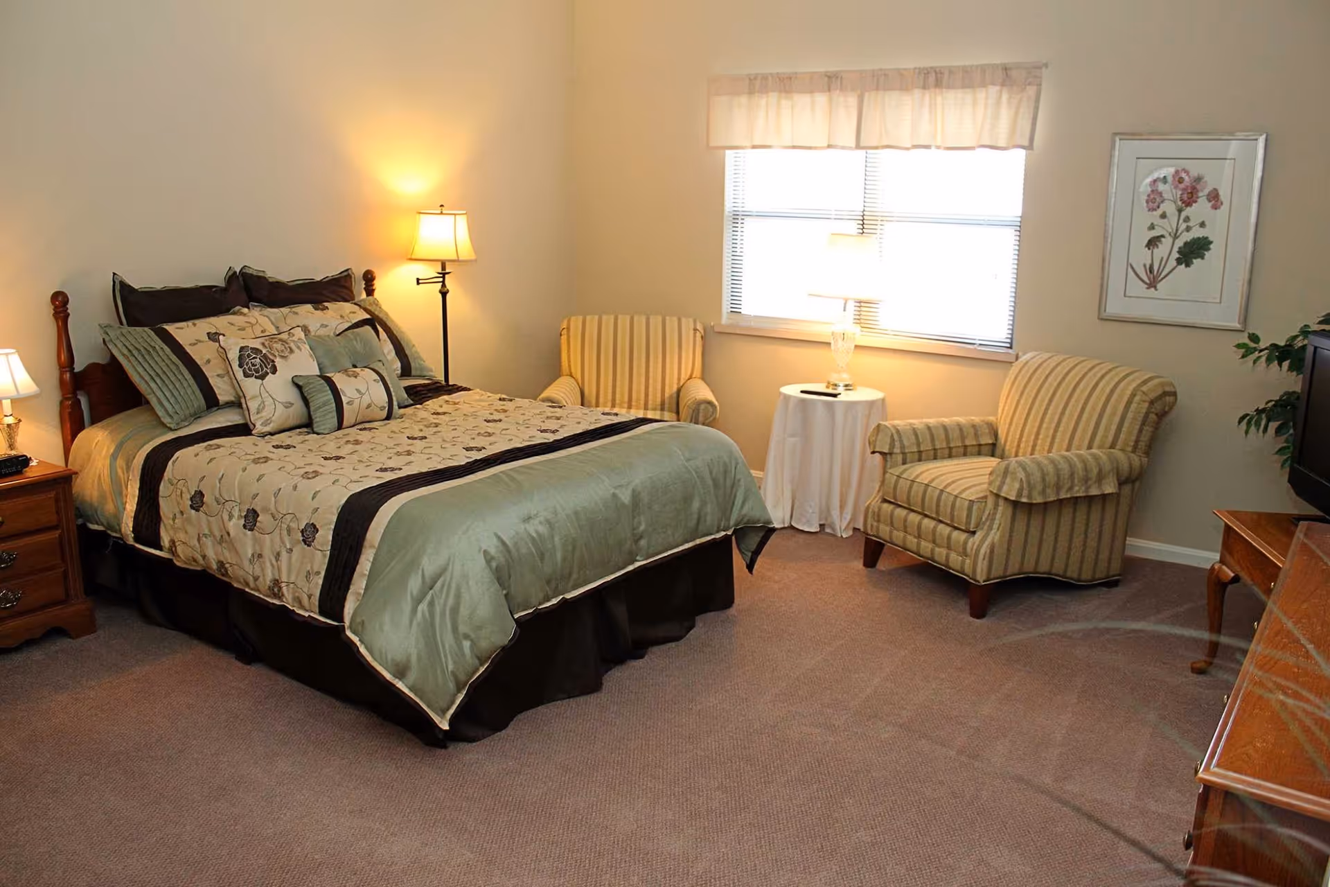 A cozy bedroom with a neatly made bed featuring floral and striped bedding. There are two striped armchairs and a small round table with a lamp between them near a window with blinds and a valance. The room also has a wooden nightstand with a lamp, a framed floral picture on the wall, and a wooden dresser with a TV on top.