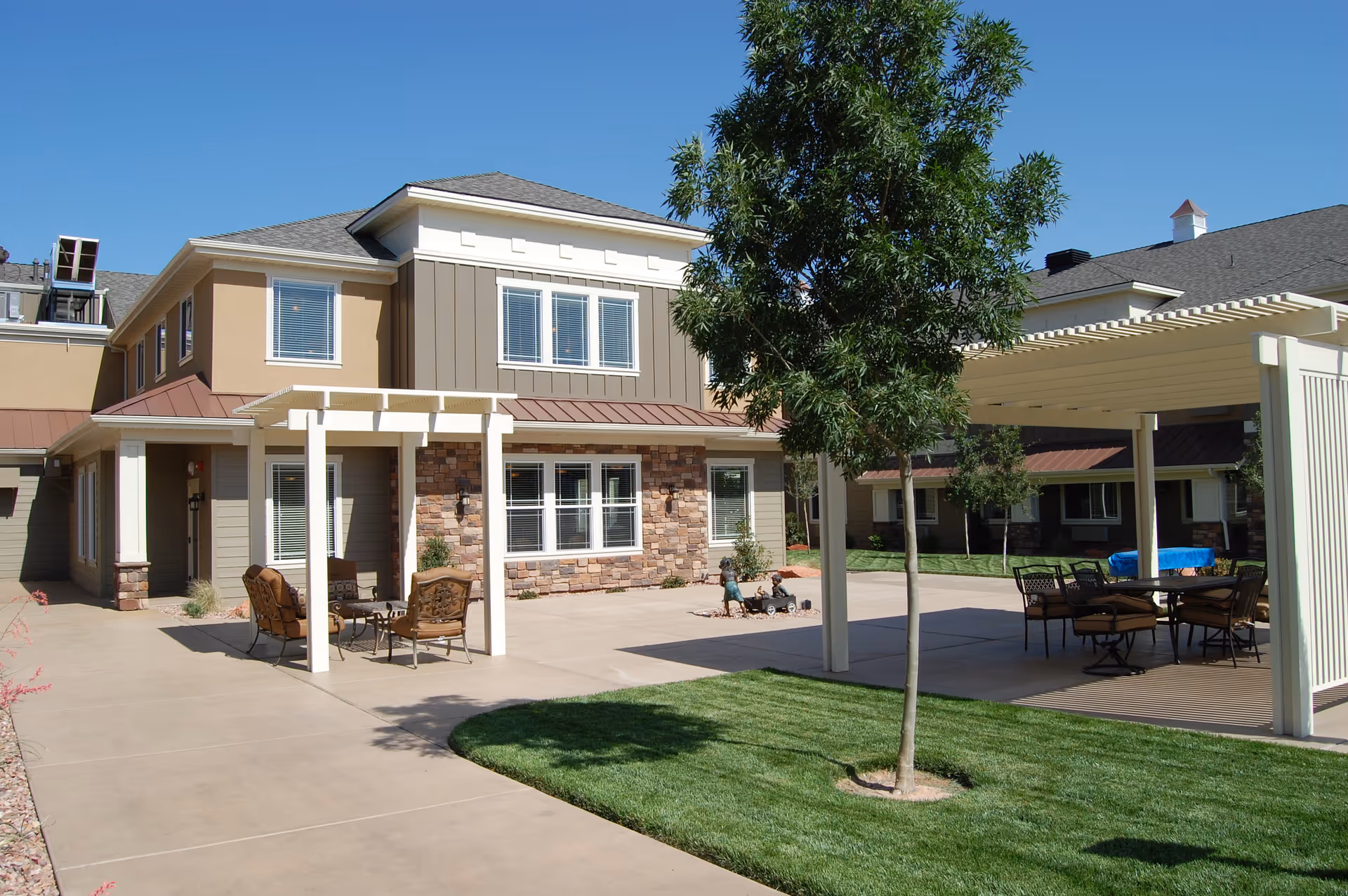 Outdoor patio area at Spring Gardens Senior Living Lindon featuring seating arrangements under pergolas, a small tree, and a two-story building with windows and stone accents in the background under a clear blue sky.