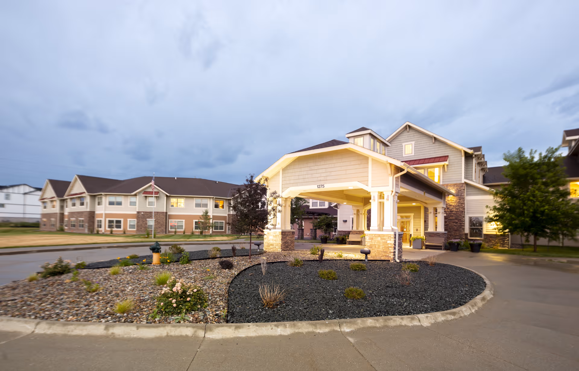 Exterior view of Independence Village of Ankeny senior living facility at dusk, showing a covered entrance with stone pillars, landscaped roundabout with plants and rocks, and multi-story residential buildings in the background under a cloudy sky.