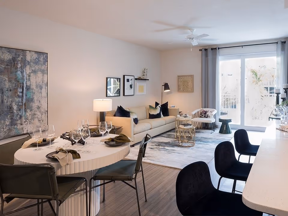 Open-plan living and dining area with a set dining table, sofa, bar stools, and sliding glass doors letting in natural light.