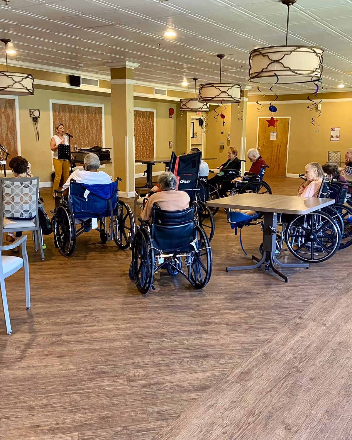 Residents in wheelchairs sit in a communal activity room watching a woman sing at a microphone.