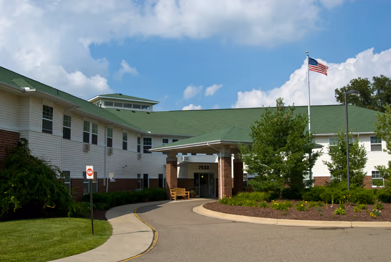 Front entrance of a two-story senior living facility with a circular driveway, green roof, and an American flag flying on a flagpole.