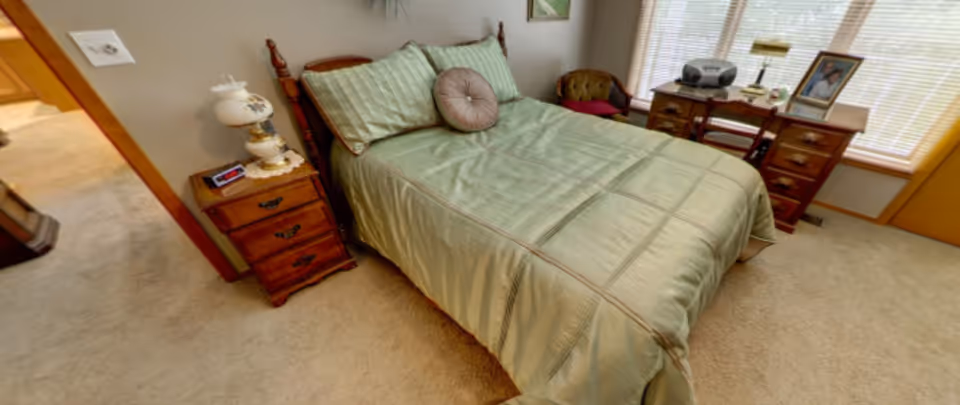 A cozy bedroom with a neatly made bed covered in a green bedspread and matching pillows. There is a wooden nightstand with a vintage lamp and an alarm clock next to the bed. A wooden desk with drawers, a chair, and a small cushioned armchair are positioned near a window with blinds, allowing natural light to fill the room. The floor is carpeted in beige.