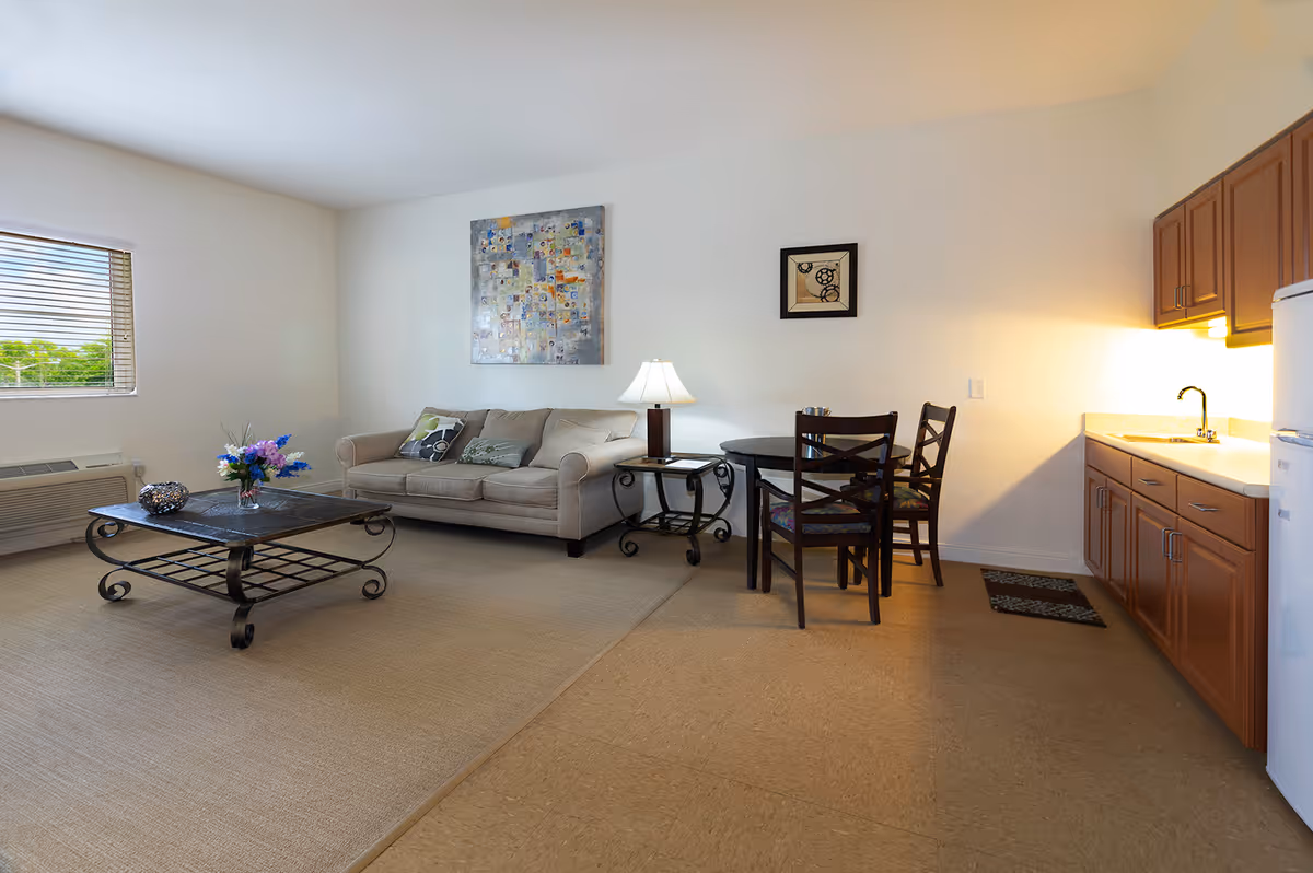 Bright living area with a beige sofa, coffee table, small dining table and chairs, and a kitchenette along the right wall.