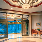 Elegant lobby area with a large chandelier hanging from a decorative ceiling. There are glass doors with intricate metal designs leading to another room with more chandeliers. To the right, there is a small seating area with two wooden chairs and a table with a flower arrangement. The floor has a polished marble design.