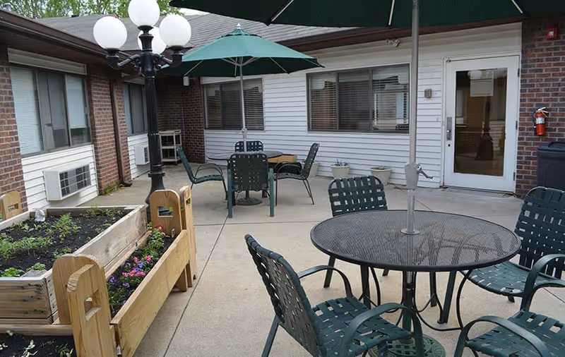 Outdoor patio area at Bethlehem Woods Nursing and Rehabilitation with round metal tables and green chairs, a large green umbrella, a wooden planter box with flowers, a black lamp post with globe lights, and a building exterior with windows and a white door.