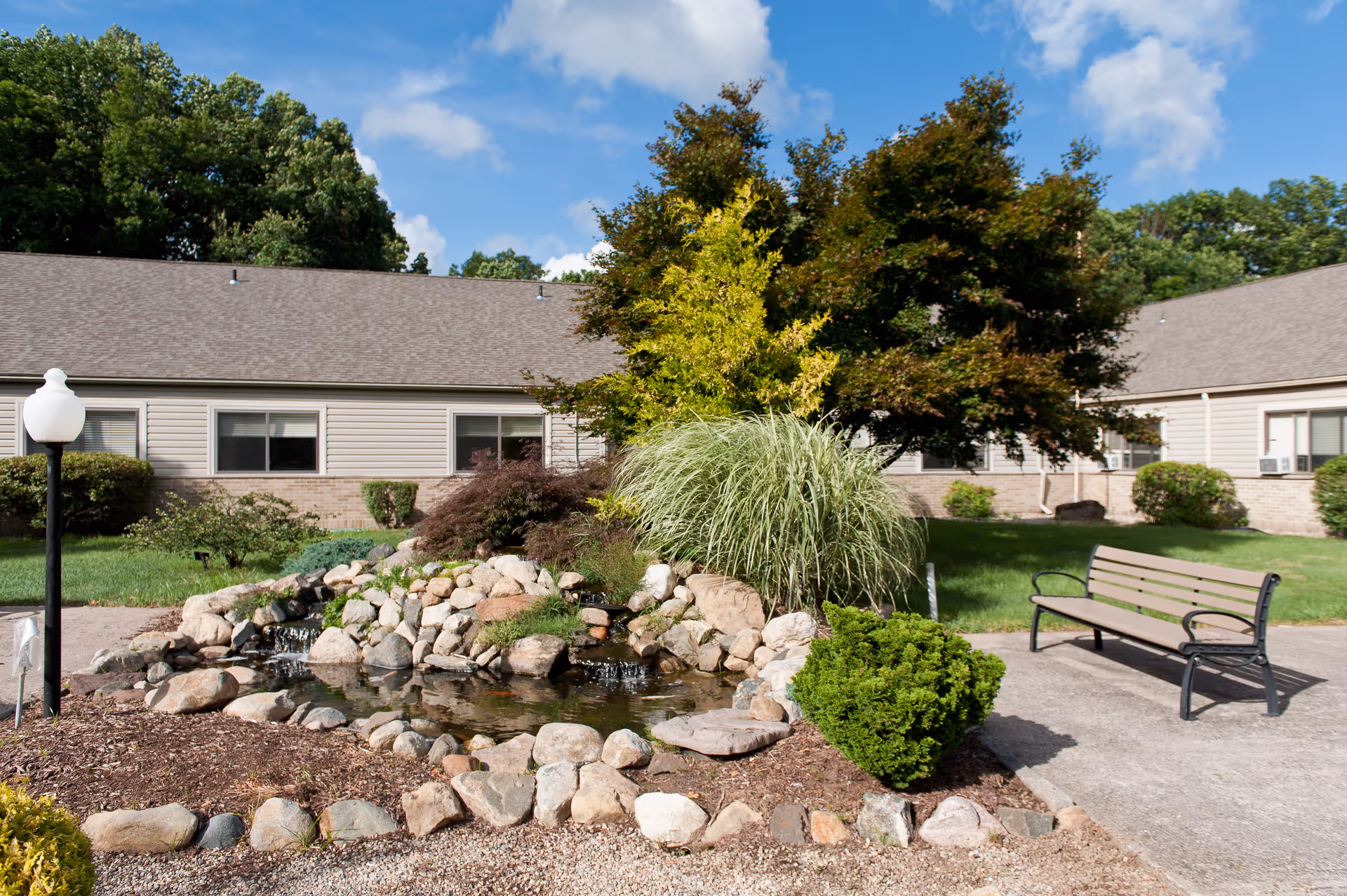 Outdoor garden area at a senior living facility with a small pond surrounded by rocks and plants, a bench on a paved path, and a building with beige siding and multiple windows in the background under a partly cloudy sky.