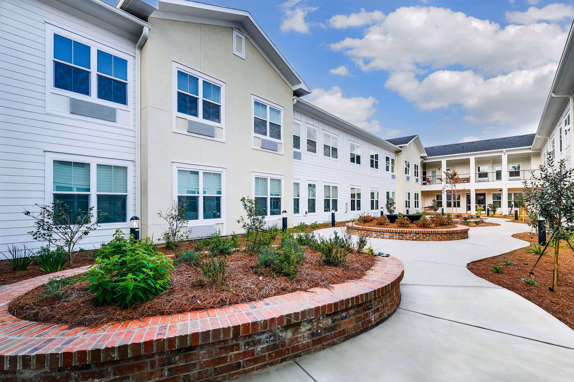 Outdoor courtyard area of The Blake at Kingsport facility featuring a paved walkway, raised brick flower beds with plants, and a two-story building with multiple windows surrounding the courtyard under a partly cloudy sky.