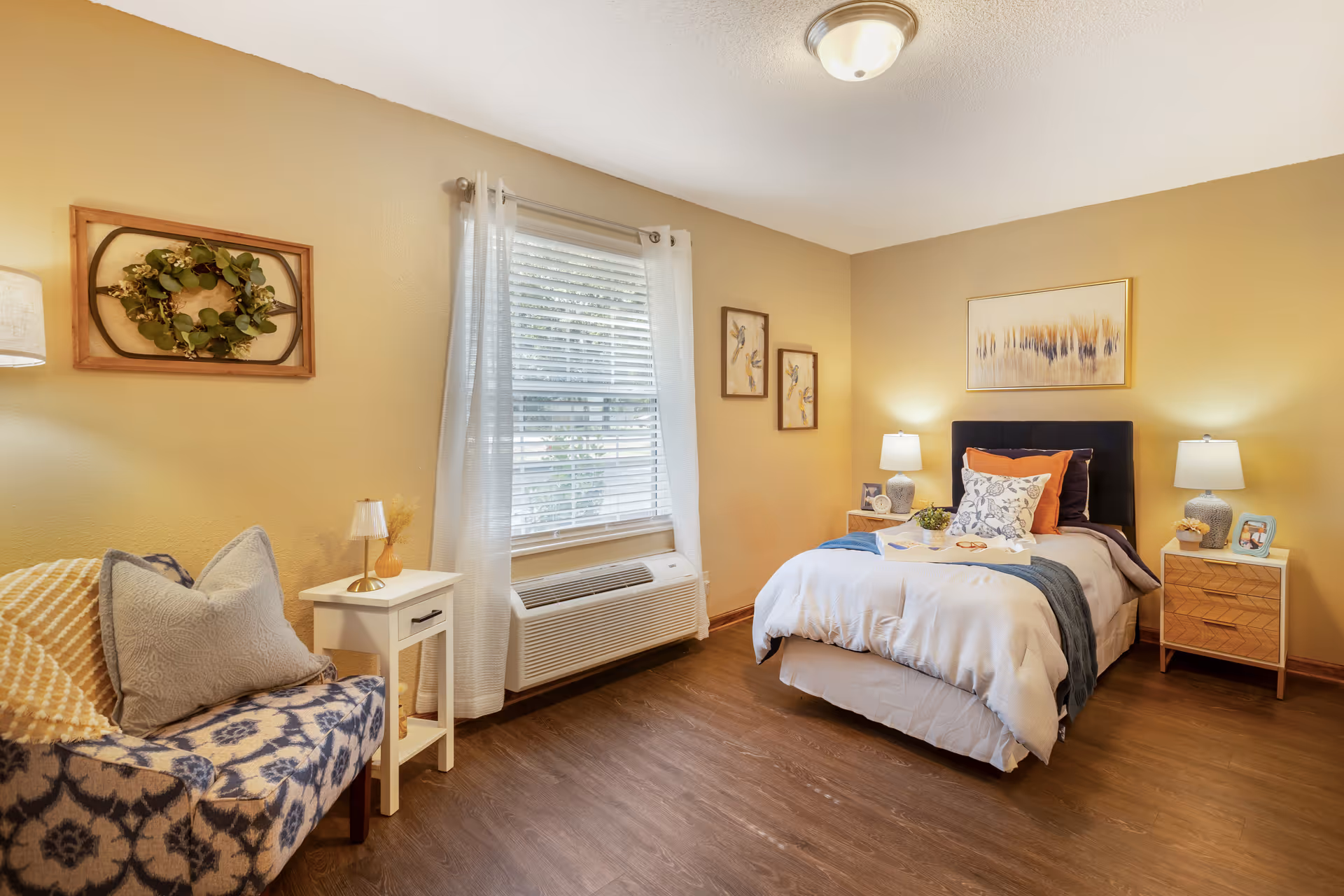 A cozy bedroom with a single bed featuring white and blue bedding, two wooden nightstands with lamps on either side, a window with white curtains, a patterned armchair with pillows, and framed artwork on the walls.