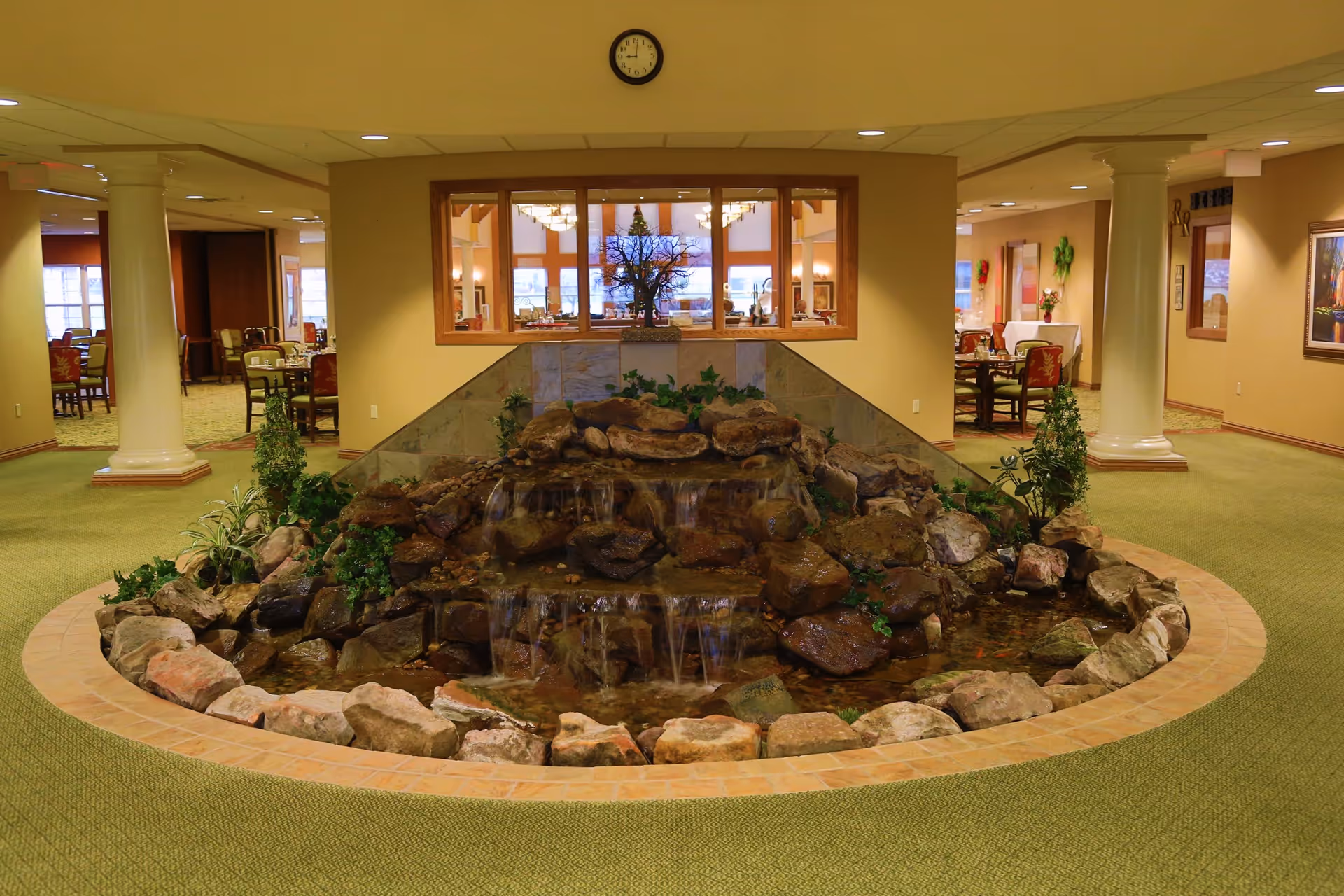Indoor water feature with rocks and small plants in the center of a carpeted room with beige walls and white columns. Behind the water feature, there is a window looking into a dining area with tables and chairs. A clock is mounted on the wall above the window.