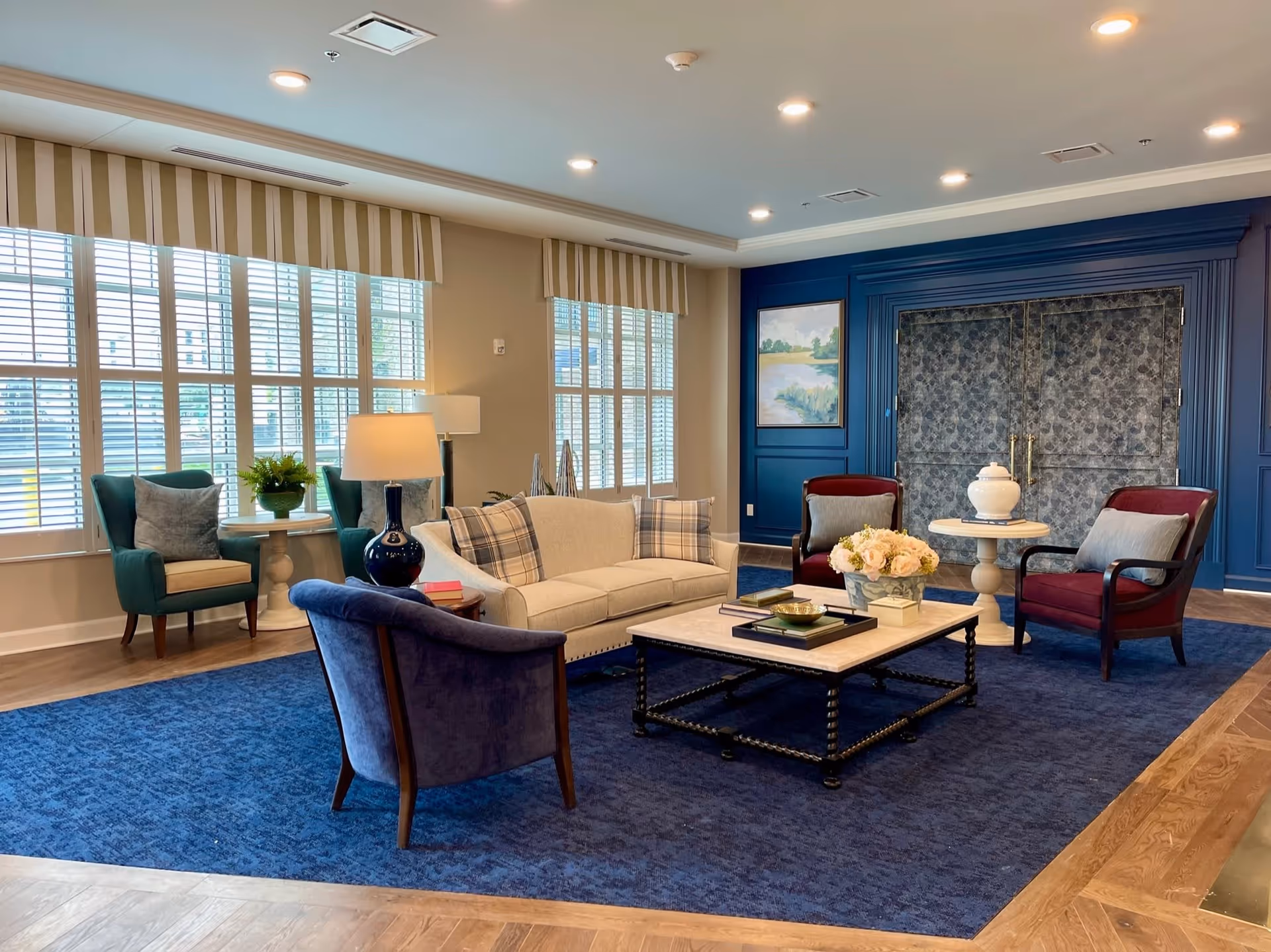 Bright communal lounge with sofas, armchairs, coffee table and large windows with shutters.