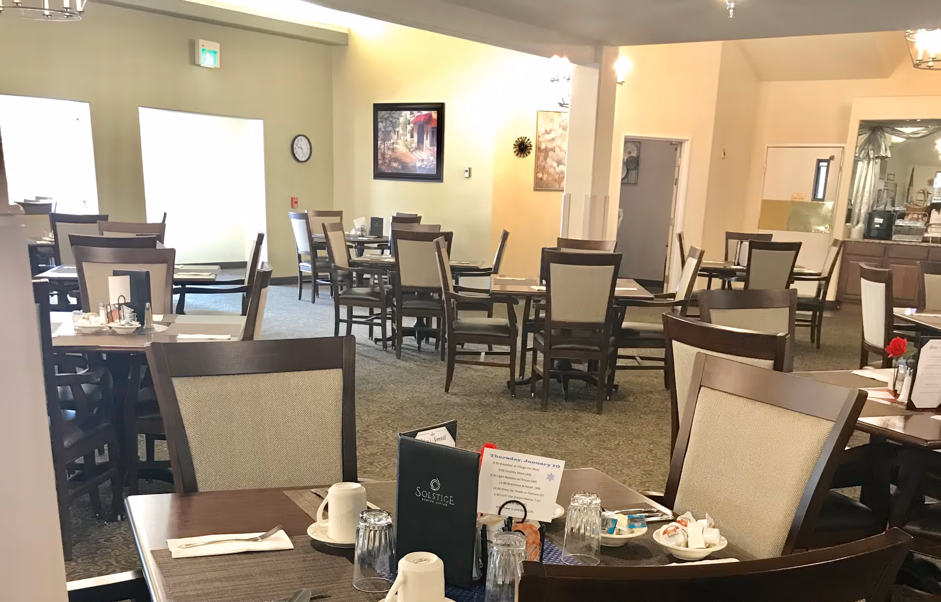 A dining room in a senior living facility with multiple tables and chairs arranged neatly. Each table is set with placemats, cups, glasses, and menus. The room has light-colored walls, carpeted floors, and framed artwork on the walls. There is a clock on one wall and a doorway leading to another room.