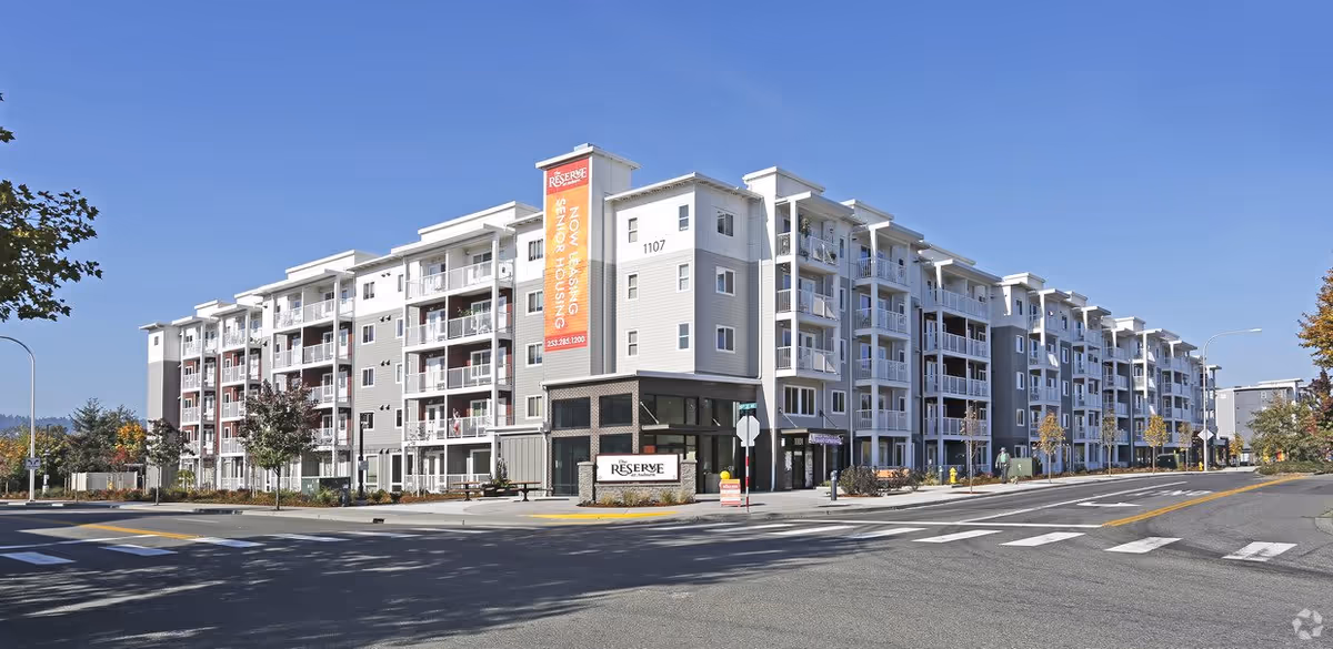 Exterior view of a modern multi-story senior living apartment building named Reserve at Auburn, with balconies and a large vertical banner advertising senior housing and leasing information. The building is situated at a street corner with crosswalks and trees lining the sidewalks under a clear blue sky.