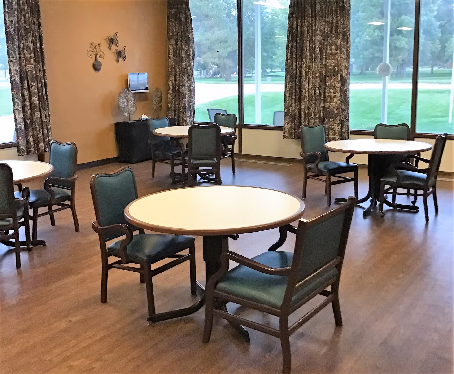 Empty dining room with round tables and green upholstered chairs and large windows overlooking a lawn.