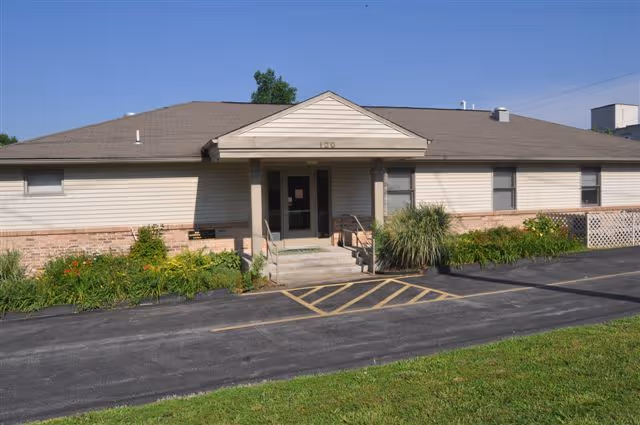 Single-story building with beige siding and a brown shingled roof, featuring a small entrance porch with steps and handrails. There are plants and shrubs along the front of the building, a paved driveway with a yellow-striped handicap parking space, and a grassy area in the foreground under a clear blue sky.
