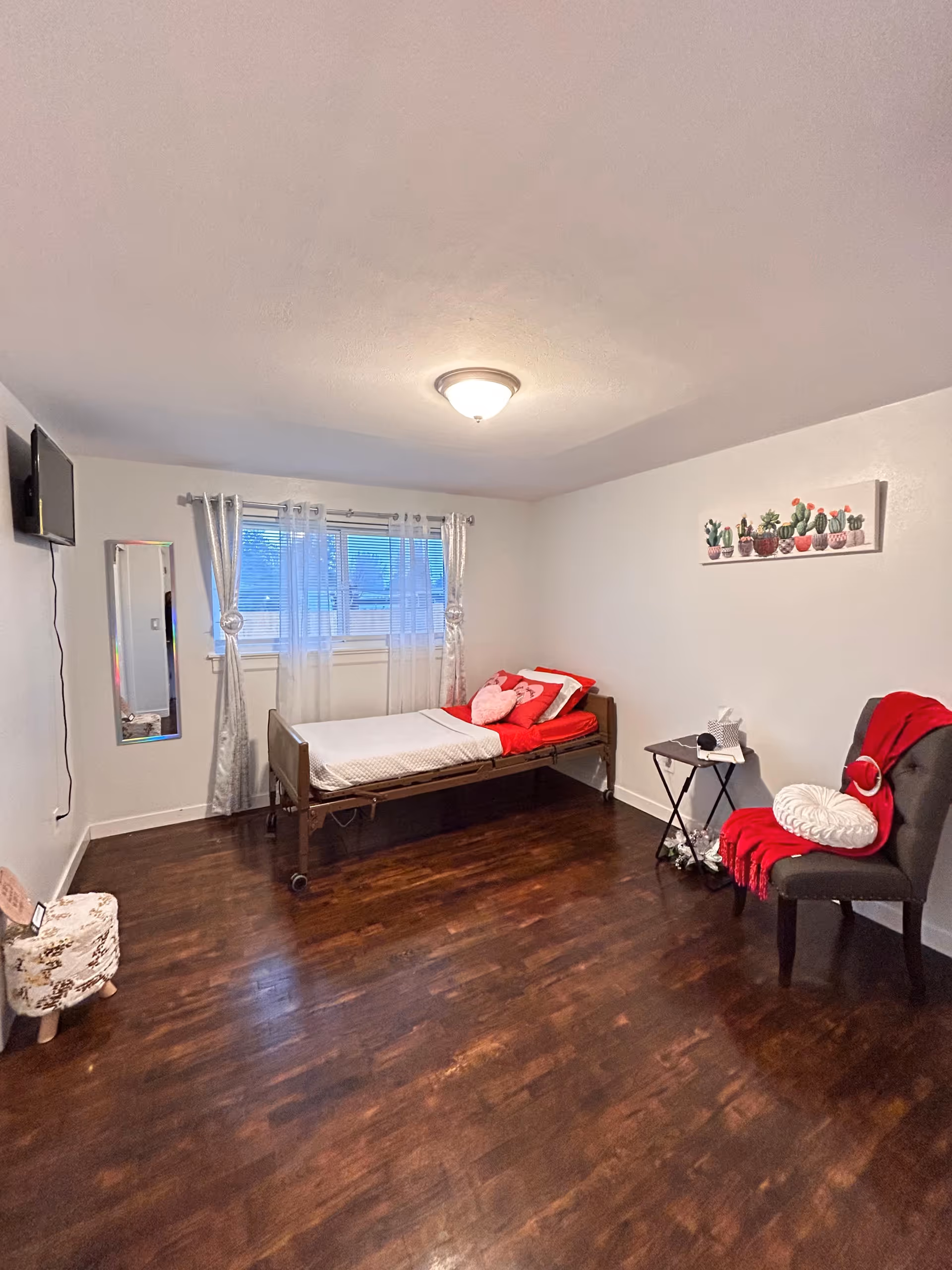 Simple furnished bedroom with a single medical-style bed against the wall, a chair with a red throw and side table, wall-mounted TV and curtained window.
