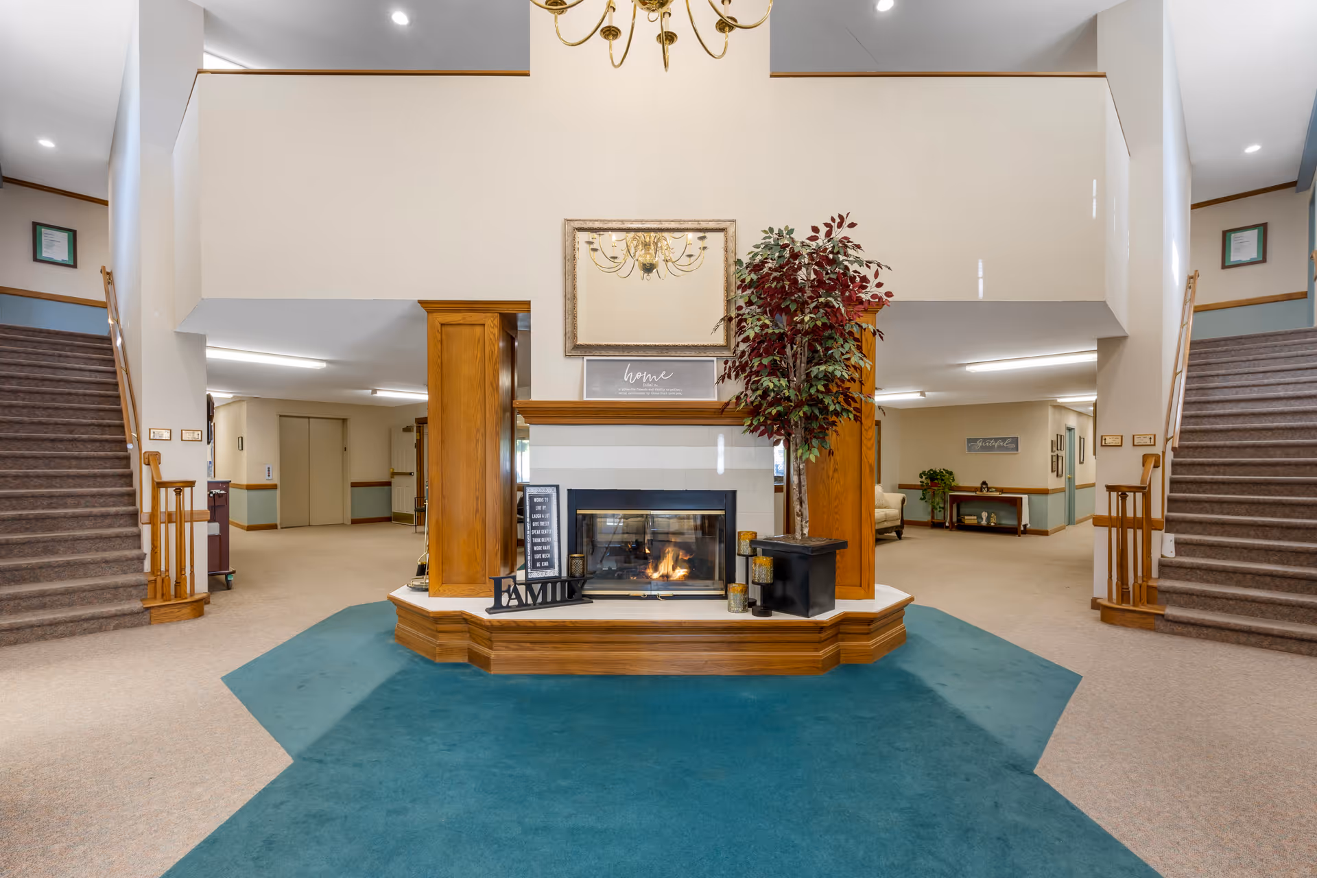 Interior view of a senior living facility lobby area with a central fireplace surrounded by wood paneling and decorative items including a potted plant and framed signs. There are two carpeted staircases on either side leading to an upper level, beige walls with wood trim, and a blue carpet in the foreground.