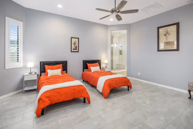 A bright bedroom with two single beds featuring orange bedspreads and white accent pillows. Each bed has a black headboard and is accompanied by a small white nightstand with a lamp. The room has light gray walls, a ceiling fan, and a window with white shutters. A doorway leads to a bathroom with a glass-enclosed shower. Two framed floral artworks hang on the walls.