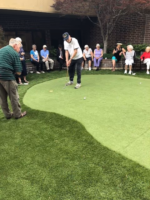 A group of elderly people sitting and standing around a small putting green outdoors. One man is putting a golf ball while others watch. The setting includes a brick wall and some trees in the background.
