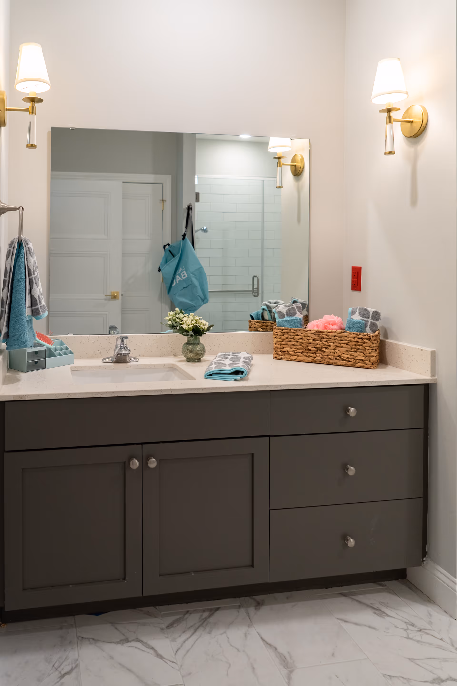 A modern bathroom vanity with a large mirror above it, two wall-mounted light fixtures on either side, a sink with a silver faucet, and a countertop decorated with a small vase of flowers, folded towels, and a wicker basket holding additional towels and a pink bath sponge. The floor is tiled with white and gray marble tiles, and a glass shower door is visible in the mirror's reflection.