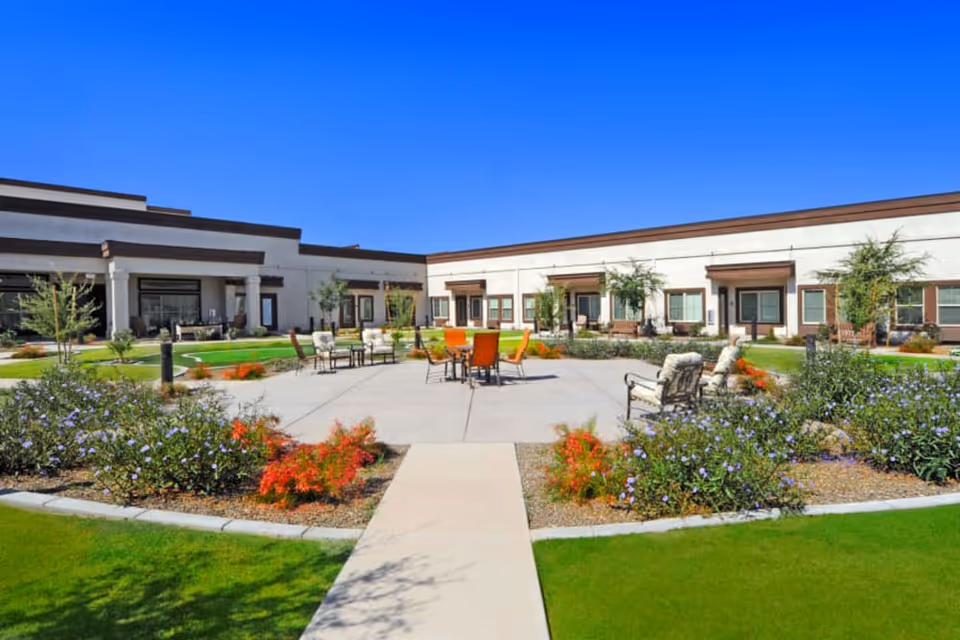 Sunny courtyard with patio seating, landscaped flower beds, and a surrounding single-story senior living building.