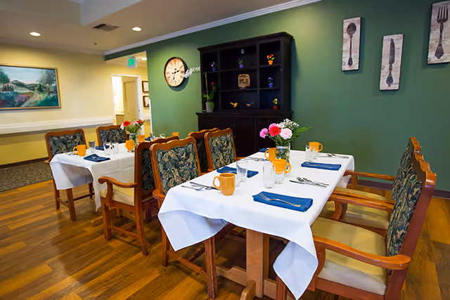 Two dining tables set with white tablecloths, blue napkins, orange mugs and floral centerpieces in a dining room with wooden chairs, hardwood floor, and a green accent wall.