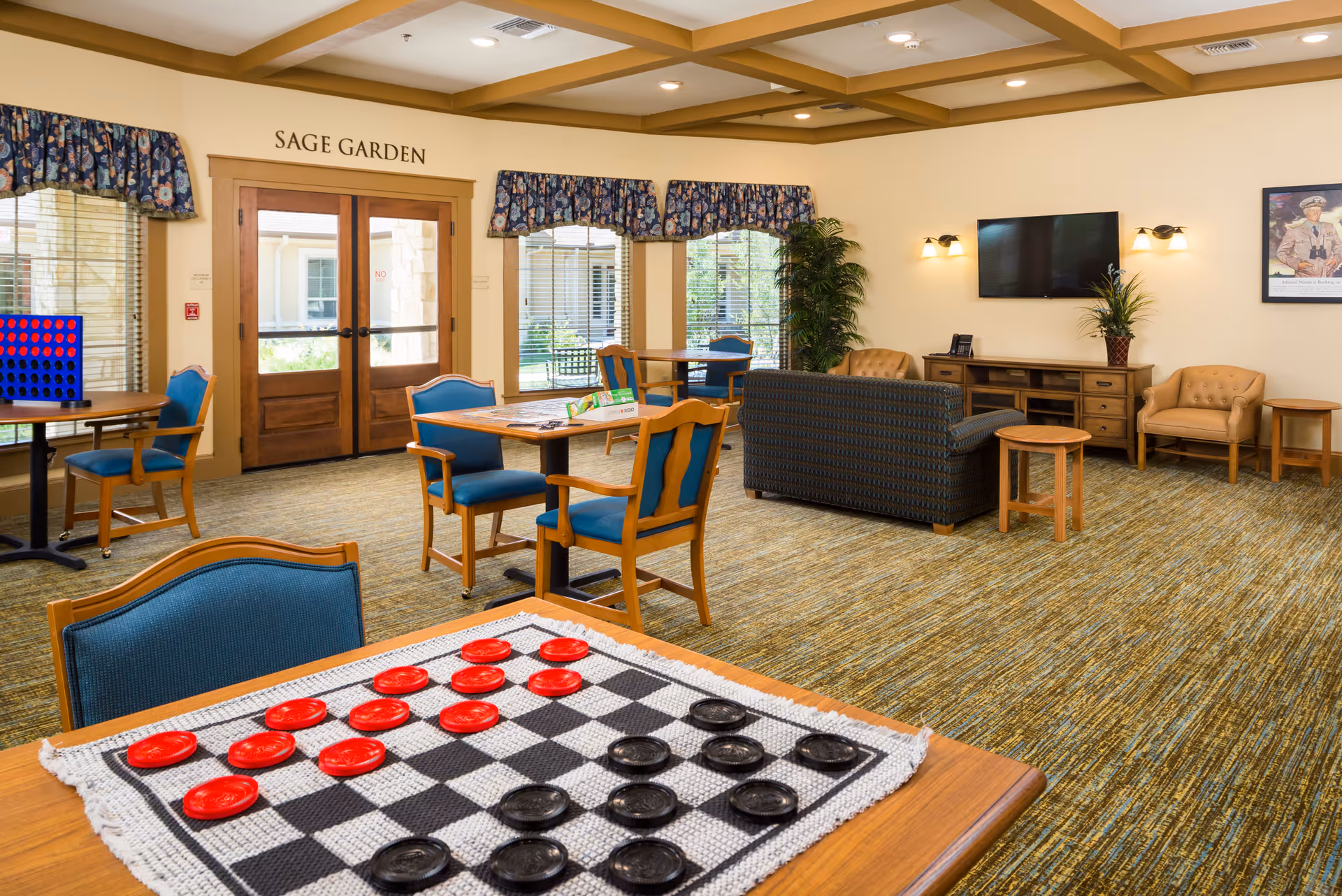 A bright and spacious common area named Sage Garden with multiple tables and chairs, a large checkers game on a table in the foreground, a blue patterned couch, two tan armchairs, a wooden TV stand with a flat-screen TV mounted above it, and large windows with floral valances letting in natural light.