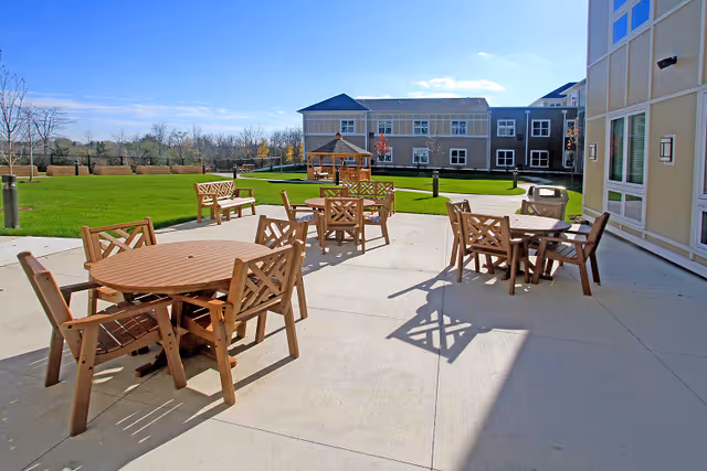 Outdoor patio area at Alto Grayslake facility with several wooden tables and chairs arranged on a concrete surface. In the background, there is a green lawn, a gazebo, benches, and a two-story building under a clear blue sky.