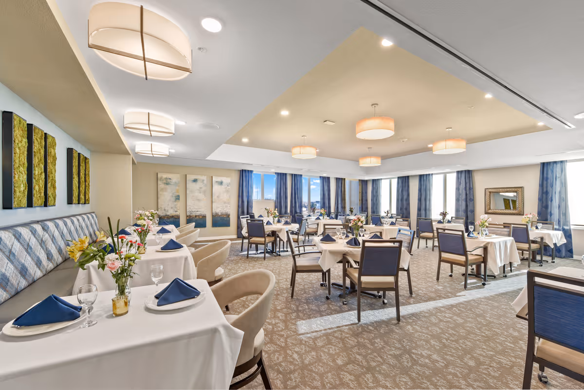 A bright and spacious dining room with multiple tables covered in white tablecloths, each set with blue napkins, plates, and glasses. The room features comfortable chairs, decorative wall art, large windows with blue curtains allowing natural light, and modern ceiling lights. Fresh flowers are placed on each table, creating an inviting atmosphere.
