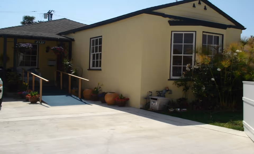 Exterior view of a single-story yellow building with a ramp leading to the entrance. There are potted plants and flowers near the entrance and windows. The building has a dark shingled roof and white-framed windows. A concrete driveway is visible in the foreground.