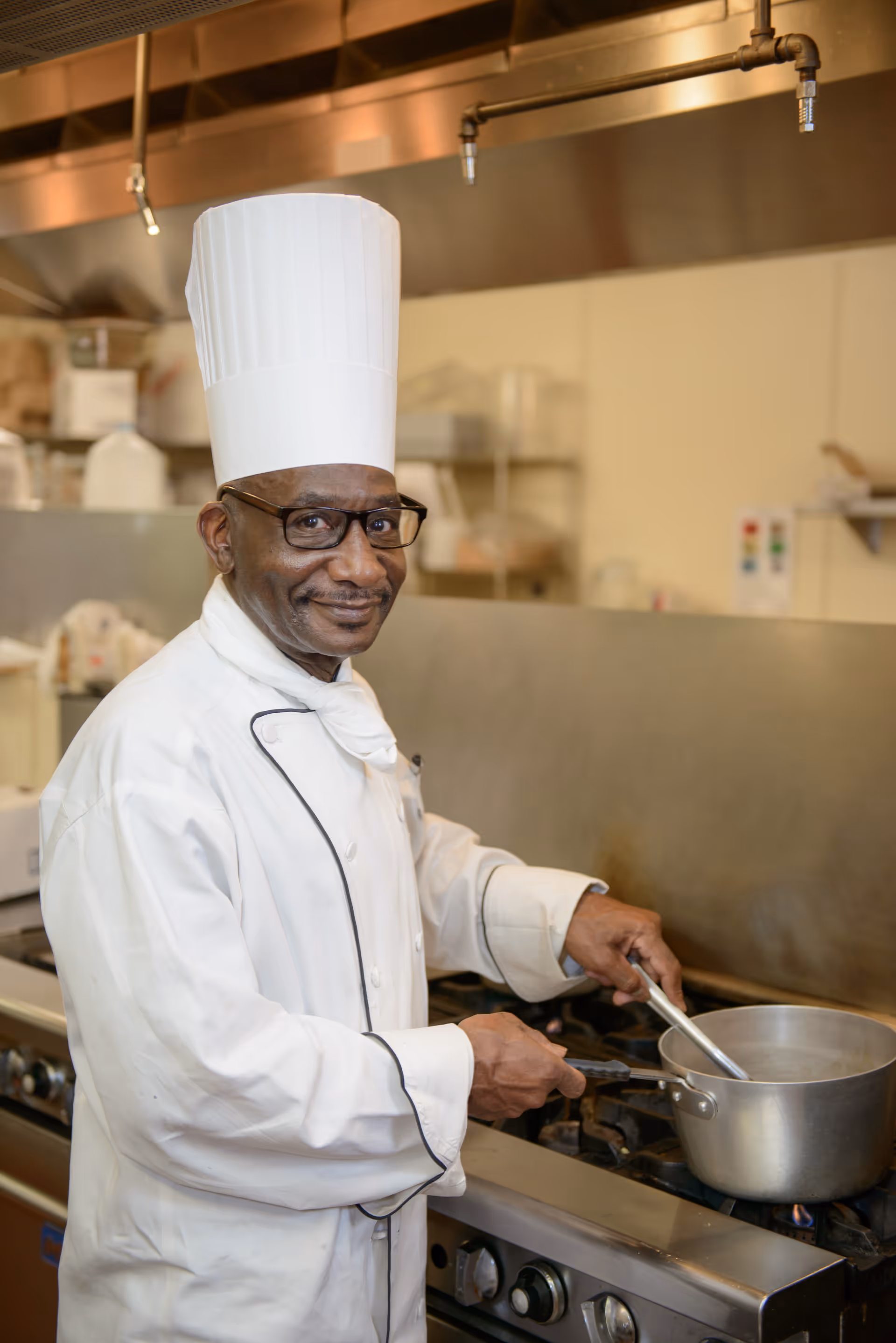 A chef wearing a white chef's coat, white neckerchief, tall white chef hat, and glasses is stirring a pot on a stove in a commercial kitchen. The chef is smiling and looking at the camera.