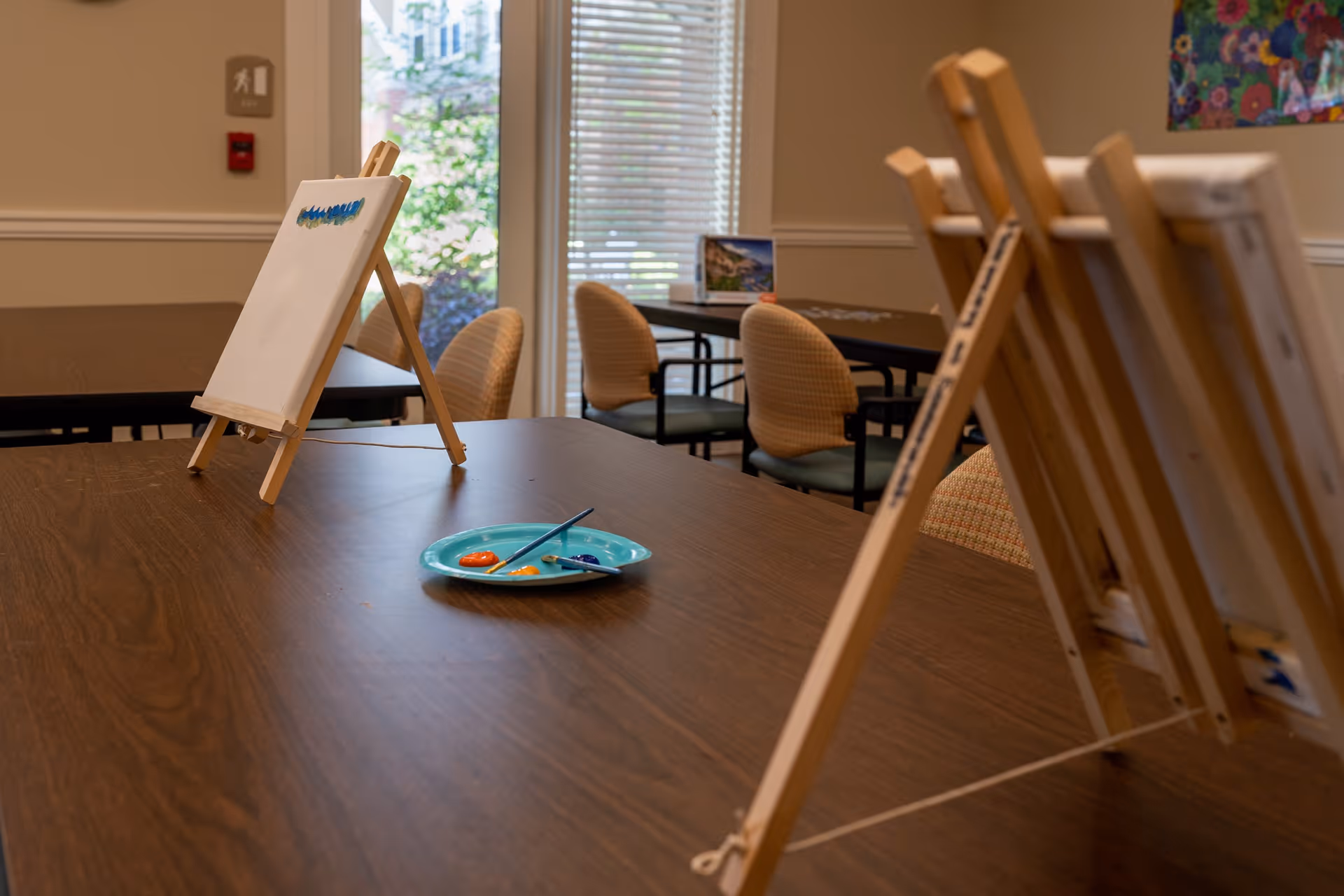A room with wooden tables and chairs arranged for painting activities. There are small easels with blank canvases and a plate with paint and brushes on the table. A window with blinds lets in natural light, and there is a fire alarm and exit sign on the wall.