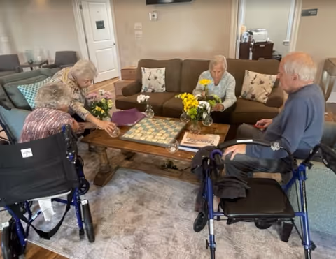 Four residents seated around a coffee table in a communal living room arranging flowers and playing a board game.