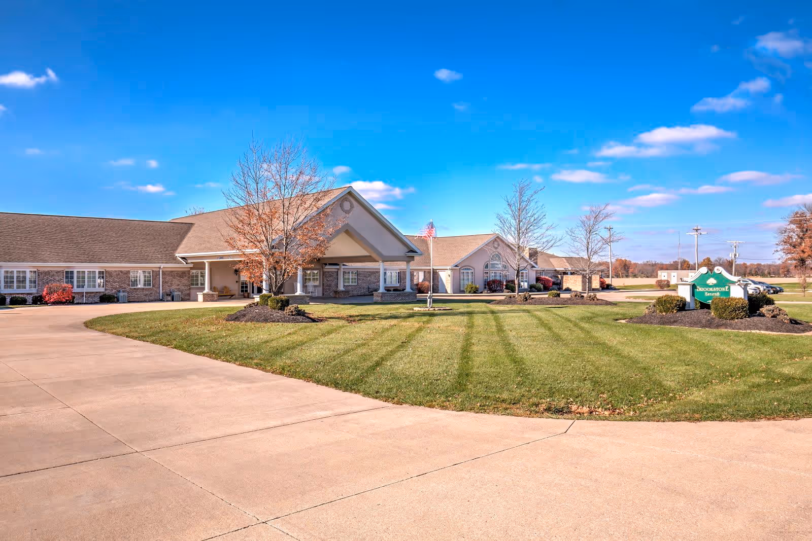 Exterior view of Brookstone Estates Of Fairfield, showing a single-story building with a covered entrance, manicured lawn, trees without leaves, and a clear blue sky.