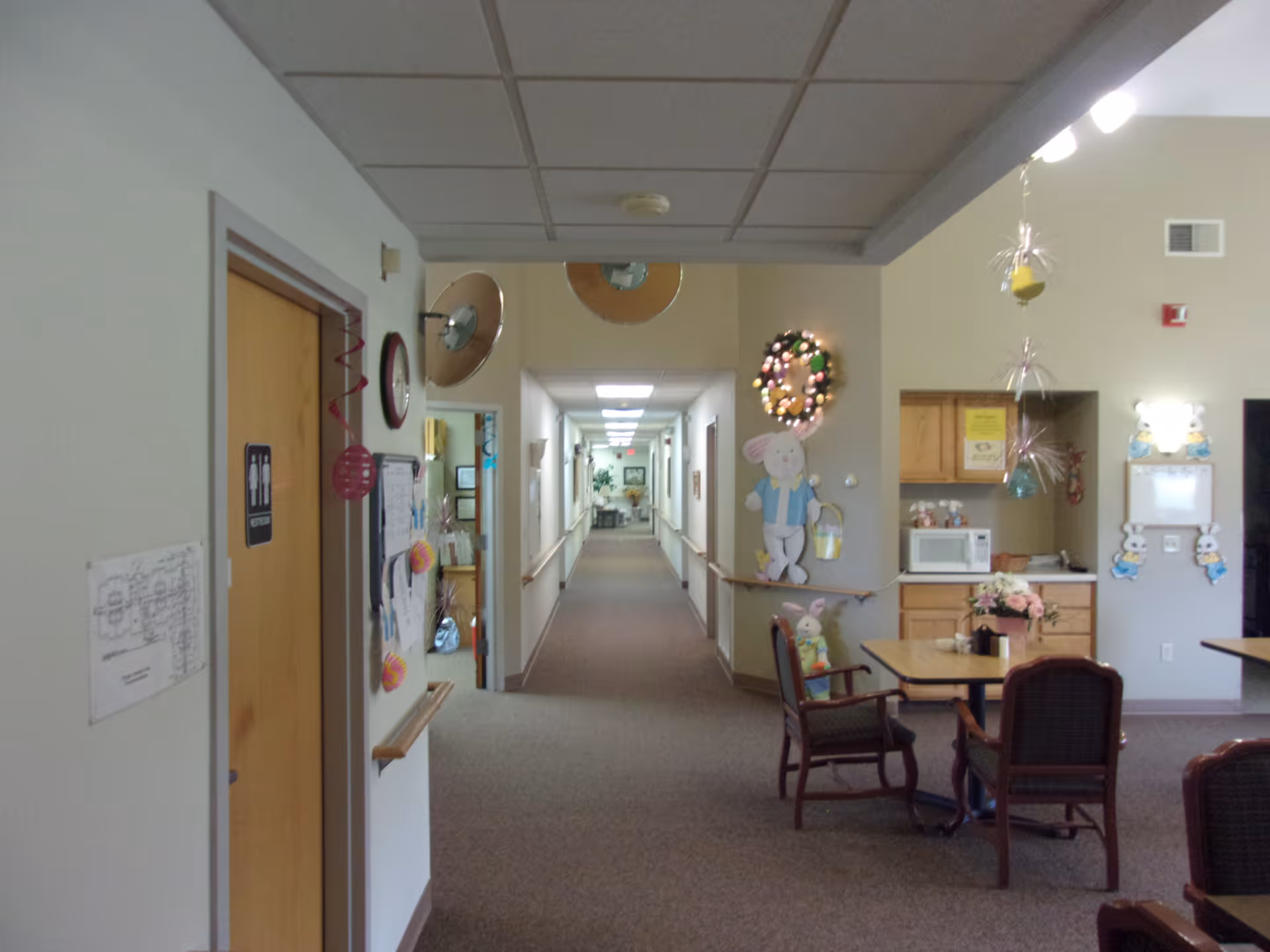 Carpeted interior hallway and common area with tables, chairs, a small kitchenette and holiday decorations in an assisted living facility.