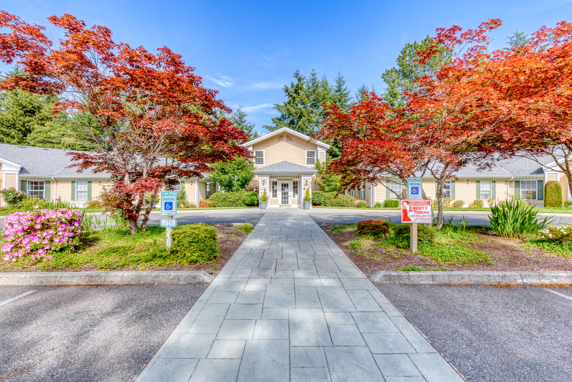Front entrance of a single-story memory care building framed by red-leafed trees, a paved walkway, and landscaped parking area.