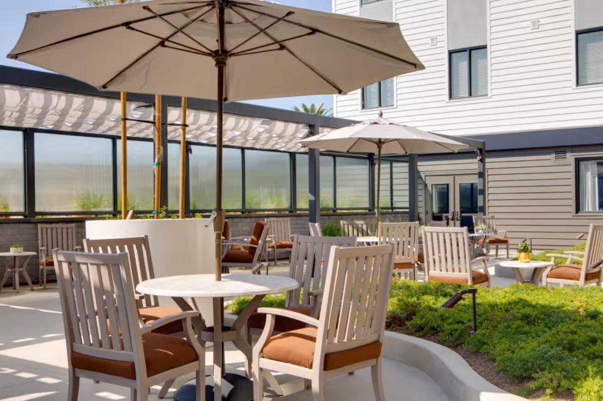 Outdoor patio area with multiple tables and chairs featuring brown cushions, large beige umbrellas providing shade, surrounded by greenery and a building with windows in the background.