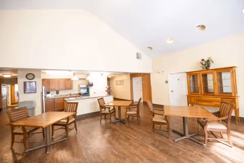 A bright dining area in a senior living facility with wooden tables and chairs arranged on a wooden floor. The room has a high ceiling with recessed lighting and a kitchen area in the background featuring wooden cabinets, a microwave, and a coffee machine. A wooden cabinet with glass doors and a flower arrangement on top is visible against the wall.