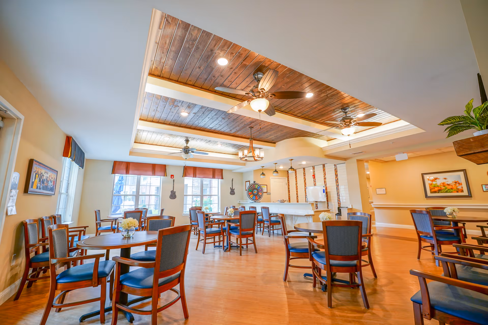 A bright and spacious dining room with multiple round wooden tables and blue cushioned chairs. The ceiling features wooden panels with ceiling fans and recessed lighting. Large windows with red valances allow natural light to fill the room. The walls are decorated with framed artwork and guitar-shaped wall hangings. The floor is wooden, and there are small flower arrangements on the tables.