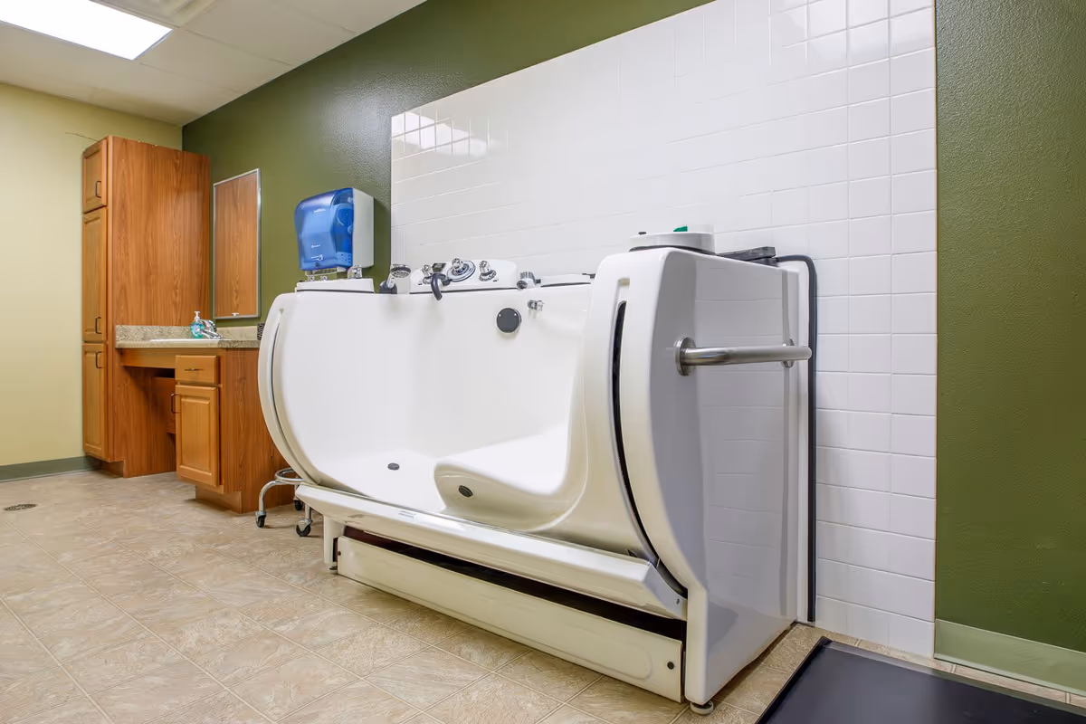 A spacious bathroom with a walk-in bathtub featuring a built-in seat and safety grab bar. The walls are painted green and beige, with white tiled backsplash behind the tub. There is a wooden cabinet and countertop with a mirror and a blue paper towel dispenser mounted on the wall.